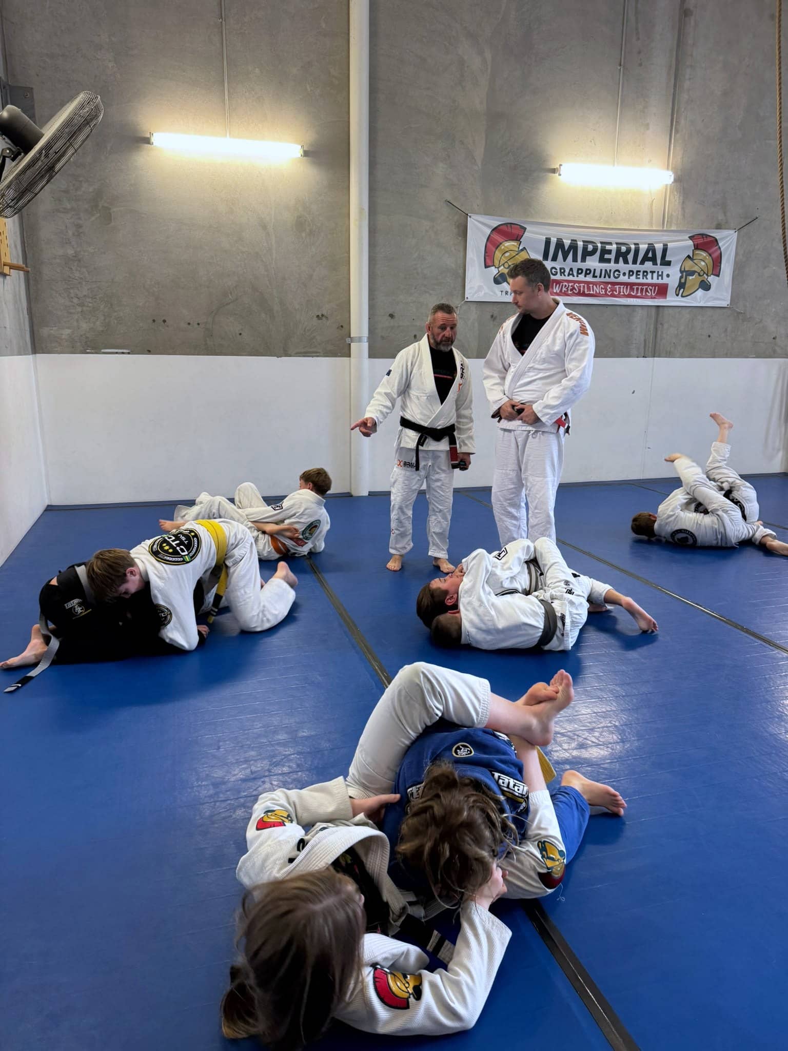 Junior members rolling in pairs across the mats while two coaches watch from the side