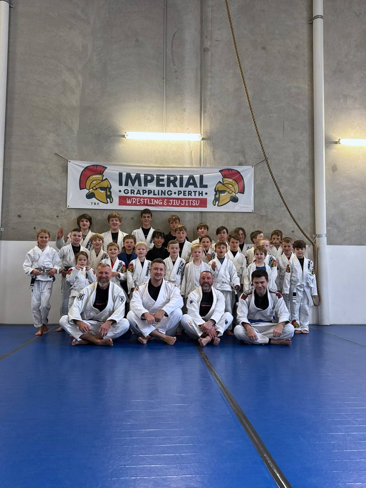 Around thirty children and teenagers in gis standing under the Imperial Grappling Perth banner, four coaches seated on the mat in front of them