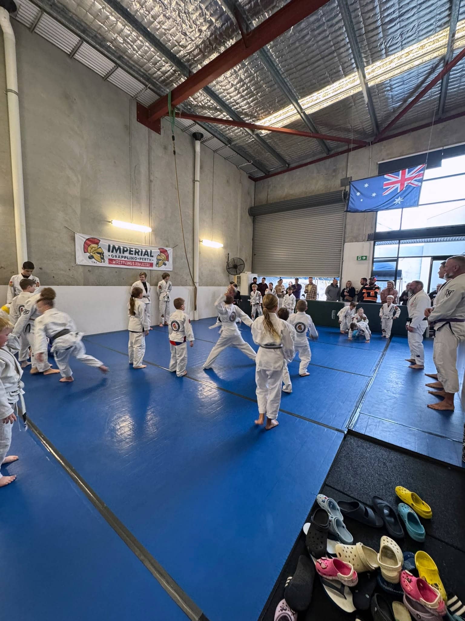 The main mat at Imperial Grappling Perth, a high warehouse space with the club banner on the wall and a class training across the floor