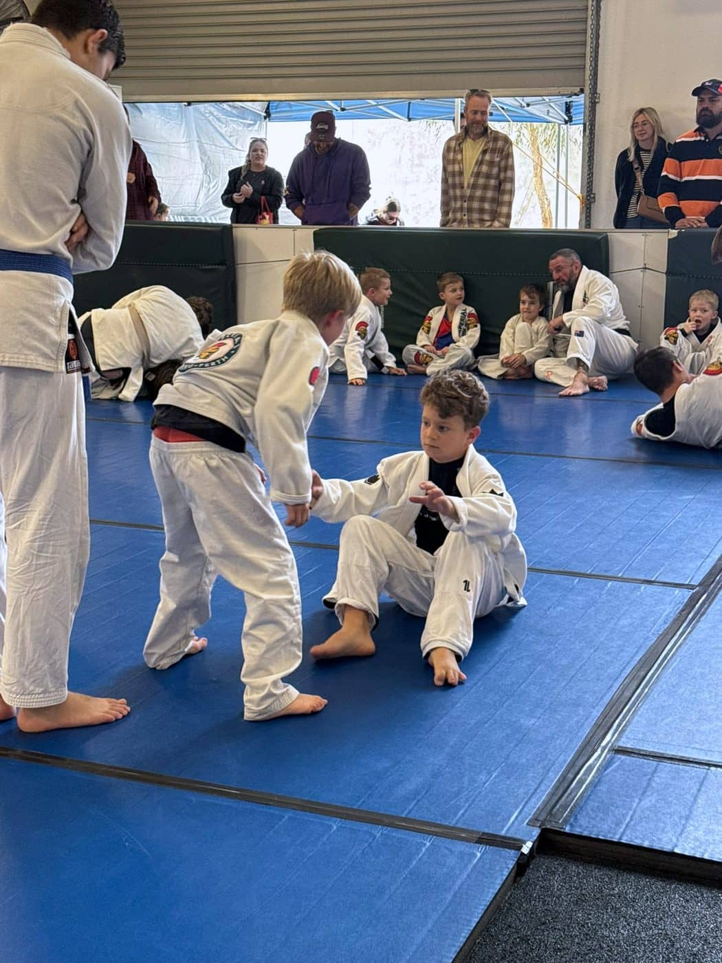 Two young members competing on the mat at an event, other kids and parents watching from the side