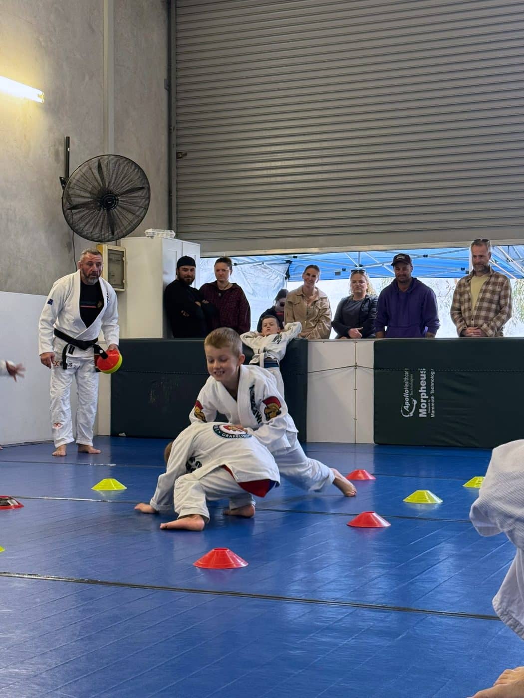 Small children working through a cone drill, one driving forward mid-effort, parents watching from behind the mat