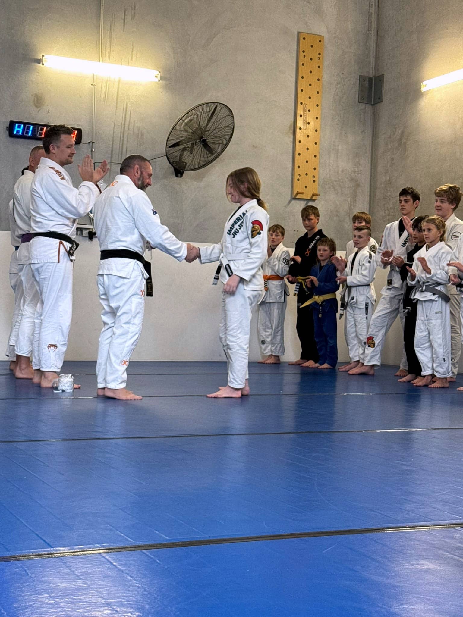 A coach shaking hands with a teenage member at a grading, the rest of the class lined up watching