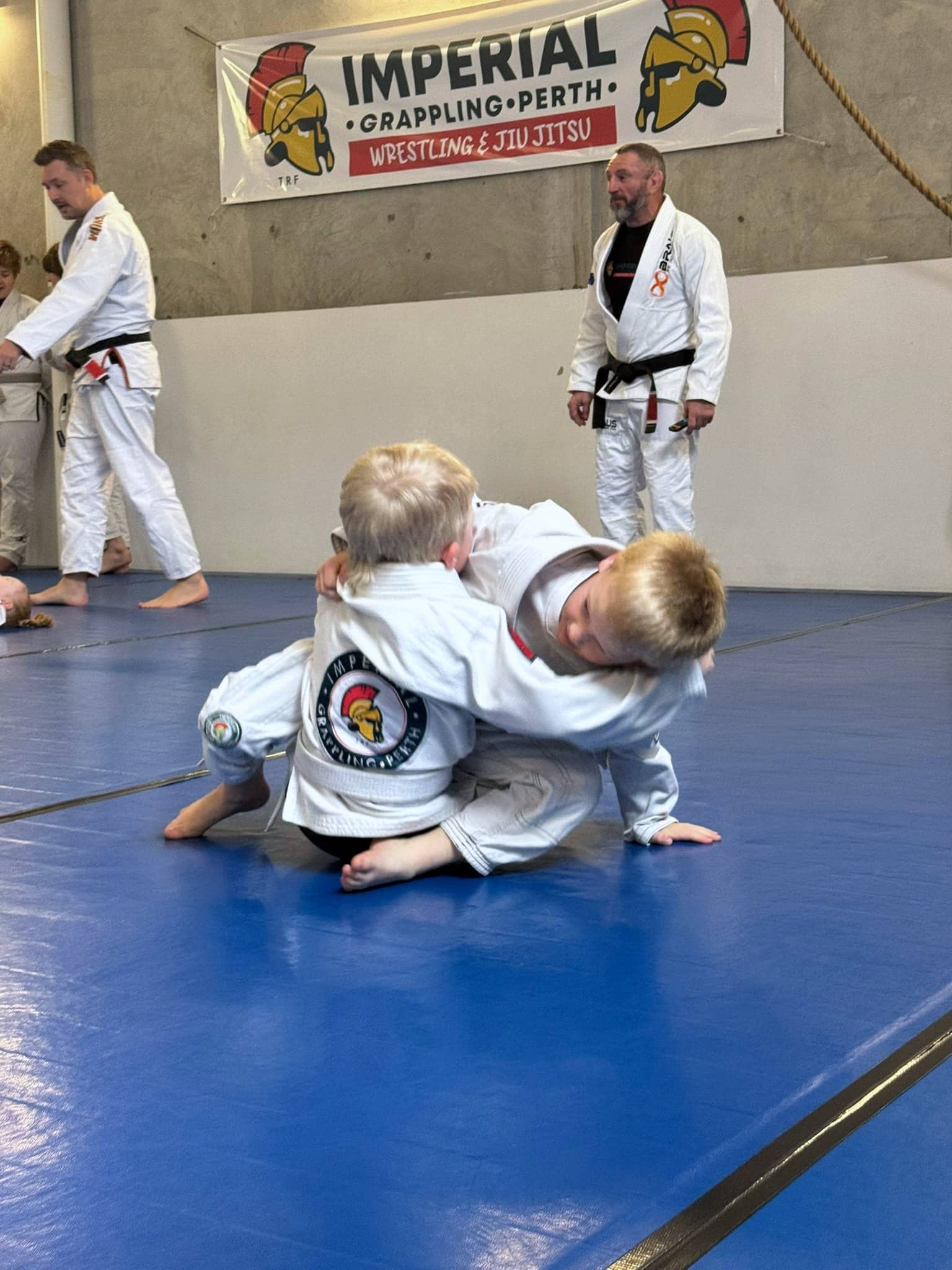 Two young children grappling on the mats at Imperial Grappling Perth while two coaches watch from beneath the club banner